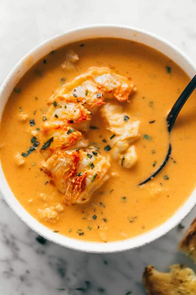 Cooked lobster flesh submerged in an orange soup in a white bowl. A black spoon is scooping some of the orange liquid in a bowl and spoon.