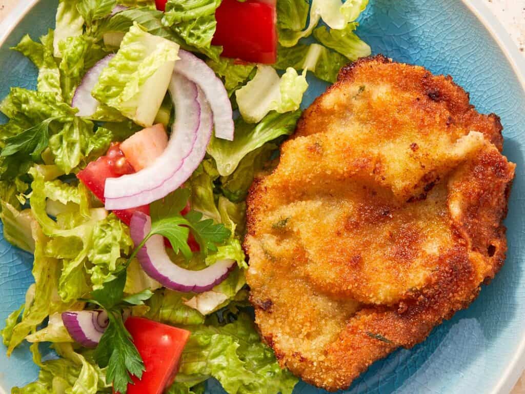 Crispy beef milanesa served with fresh tomato salad and greens on a white plate