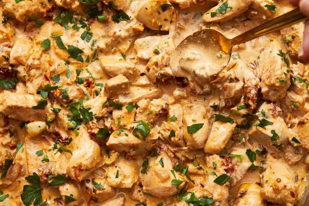 Golden spoon scooping creamy Greek Yogurt Chicken with sun-dried tomatoes and fresh parsley from a rich, velvety sauce.