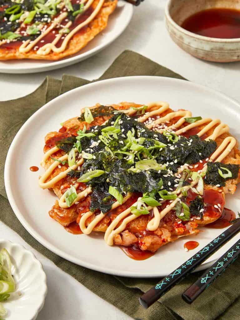 Japanese okonomiyaki pancake topped with scallions, seaweed, sesame seeds, and drizzles of mayo and sauce, served on a white plate with chopsticks.