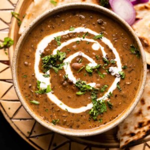 Dal Makhani served in a brown ceramic bowl with cream drizzled on top and naan and sliced onions on the side