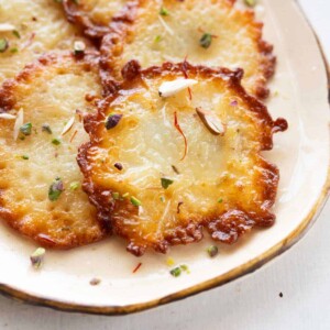 A close up shot of malpua arranged on a white plate with nuts and saffron sprinkled on top