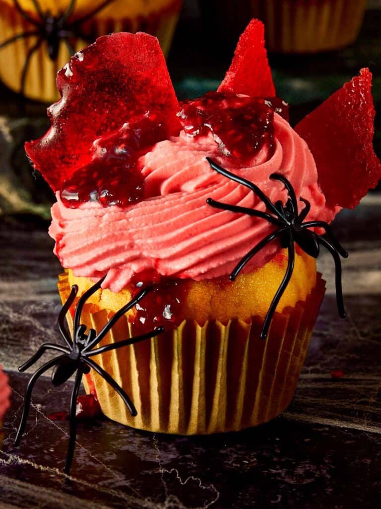 Close-up of a Halloween raspberry muffin topped with pink whipped cream, raspberry coulis, and broken glass candy shards, decorated with fake black spiders.