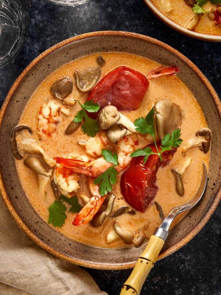 Overhead shot of creamy Thai Tom Yum Kha soup with shrimp, mushrooms, and tomatoes served in a rustic bowl with a bamboo-handled spoon and fresh cilantro on top.