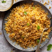 A plateful of mushroom biryani with a spoon on the side and a bowl of raita
