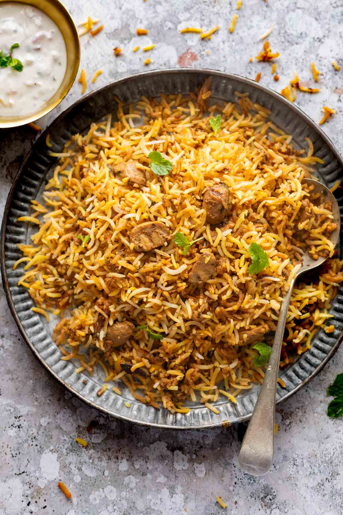 A plateful of mushroom biryani with a spoon on the side and a bowl of raita