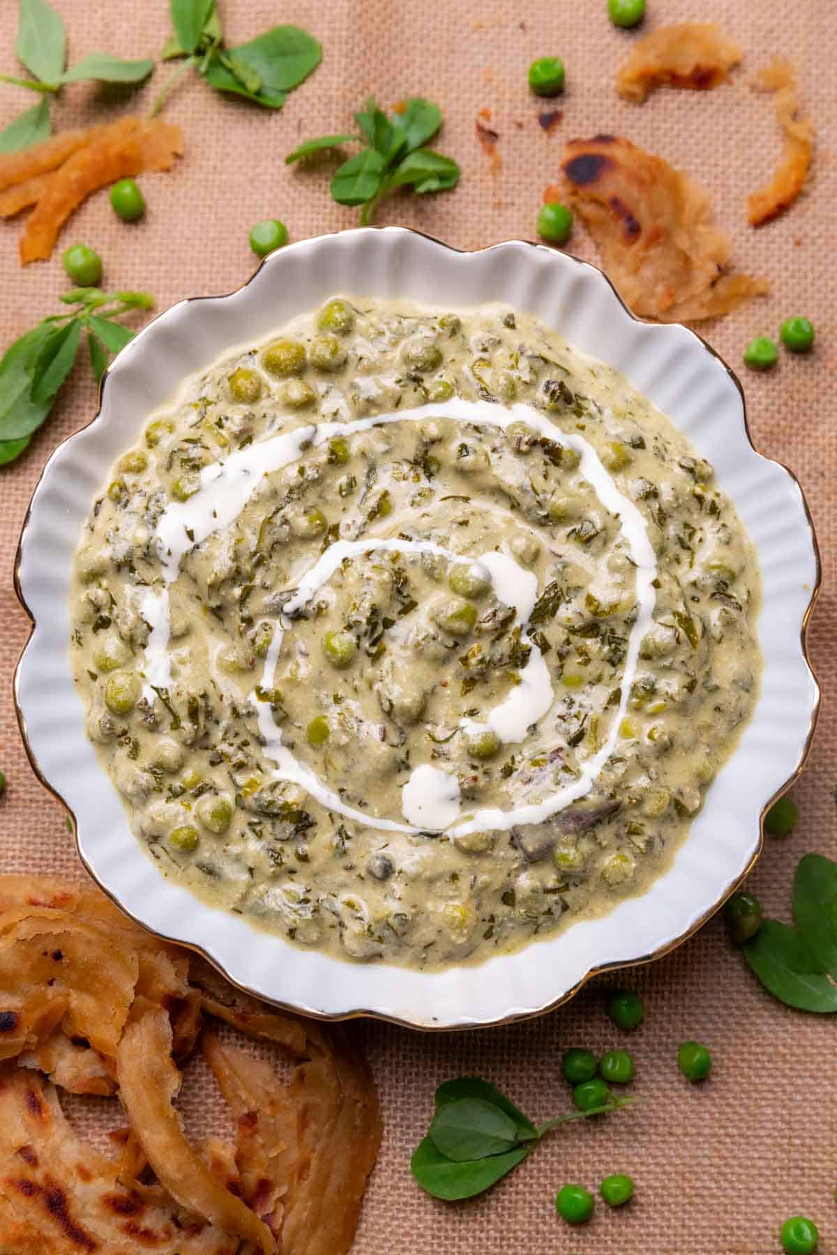 Methi Matar Malai served in a white bowl with lachha paratha, fenugreek leaves, matar etc strewn about