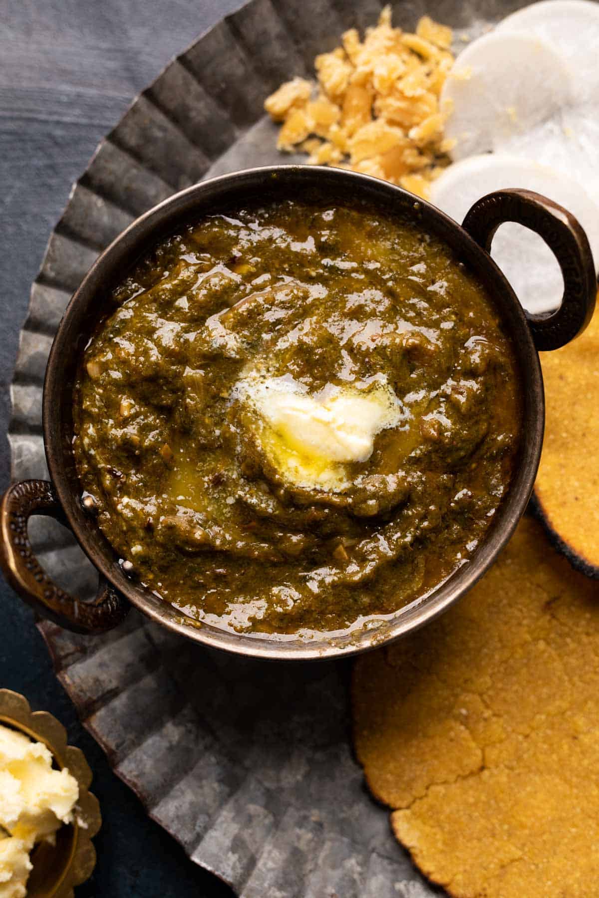 Picture of sarson ka saag served in a small copper kadhai with makki roti on the side