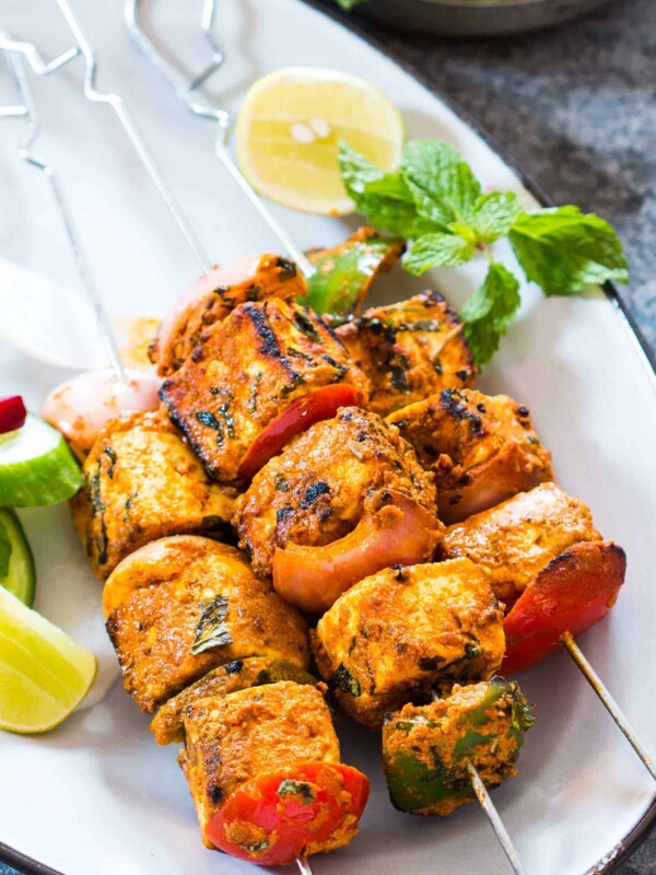 Paneer tikka skewers garnished with mint, lemon and served on a white plate.